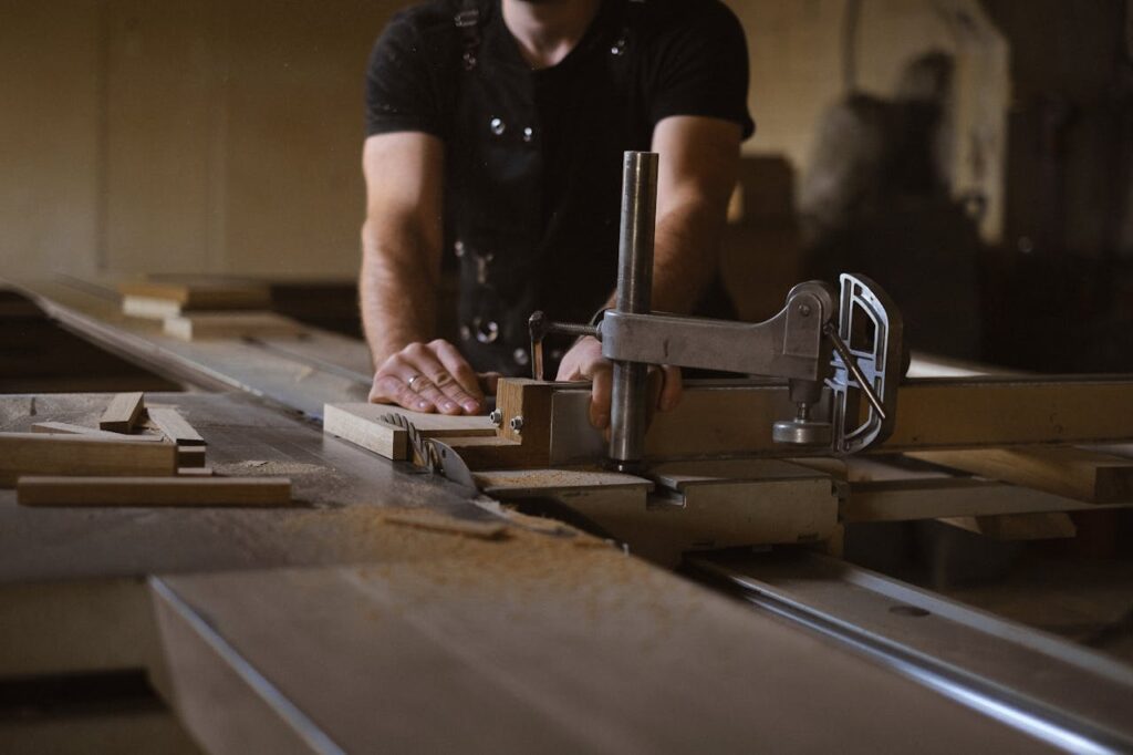 Anonymous male woodworker in apron standing at sawbench while working with wooden details in professional carpentry with stack of wood